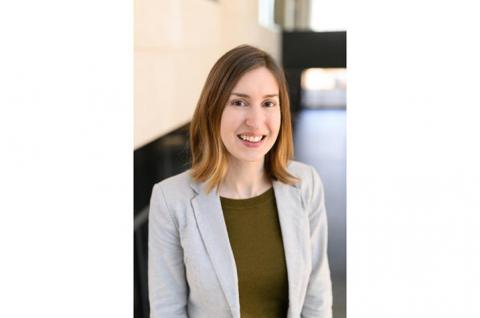 Photo of Laurie McLouth, a young white woman with straight, shoulder-length light brown hair. She's wearing a light gray blazer with an army green shirt, and smiling at the camera. Behind her is a beige wall and a window. 