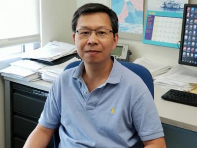 Dr. Dr. Qing-Bai She sits facing the camera with his back to his desk and computer screen. He is wearing a pale blue polo shirt and glasses. 