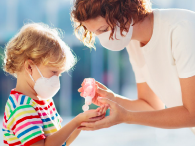 A parent in a white t-shirt squeezes hand sanitizer into the hands of a blond child wearing a rainbow striped shirt. Both people are wearing masks. 