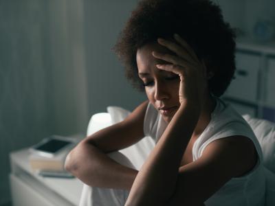 Early adult, thin Black woman with natural hair sitting in bed and holding her head with one hand.