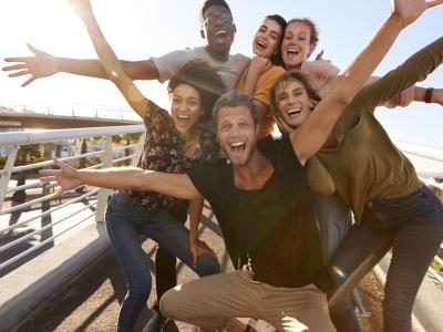 Group of smiling, multiracial young adults gather together with outstretched arms. They're wearing short sleeves and are standing on a pedestrian bridge on a sunny day. 