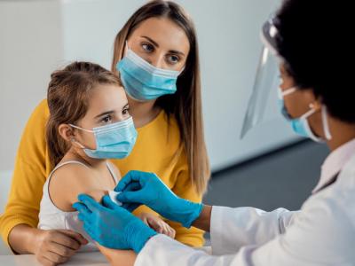 A Black female doctor applies a bandaid to the arm of a white little girl who's sitting with her mother. We see the side and back of the doctor's head; she has short hair and is wearing a white coat, blue gloves, and blue surgical mask. The little girl has long medium-brown hair pulled back in a pony tail and is wearing a white tank top. Her mother sits with her arm around the little girl; she has long medium brown hair and is wearing a yellow long-sleeve shirt. 