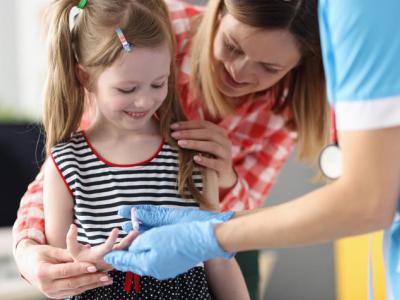Stock image showing a child (female, white, with long hair pulled back in pig tails)  checking their blood sugar with a finger prick. Their mother and a health care provider assist. 