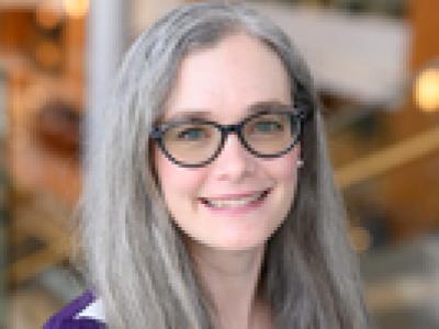 Photo of Dr. Amy Meadows, a white woman with long, straight gray hair and brown glasses. She's wearing a long sleeve dark purple shirt and smiling at the camera. 