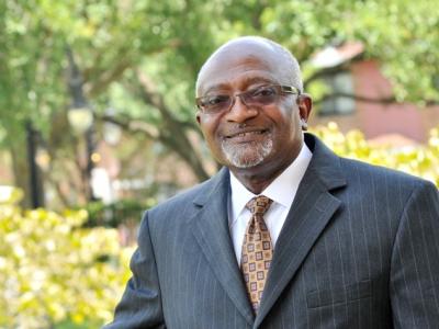 Dr. Robert Bullard in a dark gray suit stands smiling with trees out of focus behind him. 