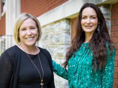 Waist-up photo of UK researchers Christal Badour, left, and Mairead Moloney standing outside a brick and stone building. Badour is on the left, a white woman with straight blond hair just past her chin. Moloney is on the right, a white woman with long, dark, wavy hair, wearing a teal blouse with dark and light blue dots. 