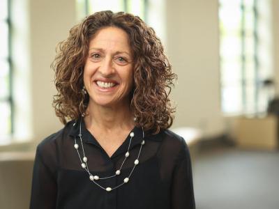 Torso headshot of Nancy Schoenberg, a middle-aged white woman with shoulder length curly hair. She's smiling at the camera, wearing a black collard blouse and a double drape necklace with intermittent pearls. 