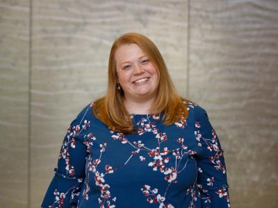 Allison Gibson, PhD, a white woman in her 30s, stands in front of a light gray wall, smiling at the camera. She has strawberry blond, straight hair past her shoulders, and she's wearing a long-sleeved blue dress with cherry blossoms. 