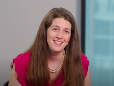 Dr. Mary Sheppard, a white woman in her mid 30s, smiles at the camera; she has long, thick, straight, light-brown hair and is wearing a pink short-sleeved blouse with a double strand of pearls. She's sitting in front of a light gray wall and a large window. 