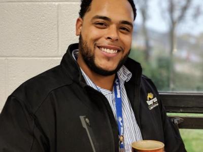 Robert Bell, a Black man in his late twenties, smiles at the camera. He's wearing a light blue striped collared shirt and a black jacket that's upzipped. He's standing in front of a window and holding a coffee mug. 