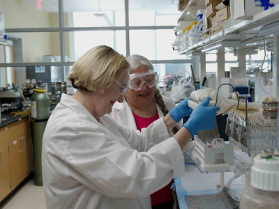 Two white women in their 50s or 60s, wearing white lab coats, blue medical gloves, and goggles, stand at the bench of a scientific laboratory; they're working with test tubes and equipment. 