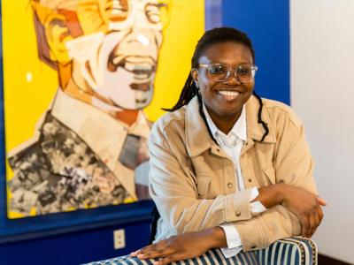 Princess Magor Agbozo, an undergraduate student in public health, stands in front of a large, bright yellow portrait collage of Lyman T.  Johnson. Agbozo is a young Black woman wearing a beige corduroy jacket with a white collared shirt underneath. She's wearing clear-rimmed glasses, her long twists pulled back in a ponytail, and she's smiling at the camera while leaning on the back of a striped wingback chair. 