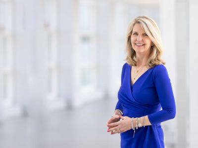 Dr. Susan Smyth, a white woman with blond, shoulder length hair, stands in a  white room with floor to ceiling windows behind her. She's wearing a royal blue v-neck dress and several bracelets. 