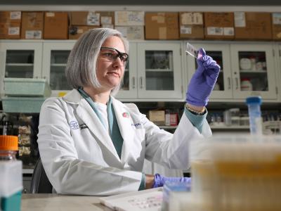 Jill Roberts, PhD, sits at a workbench in her lab. She has gray hair cut below her chin and she's wearing a white medical coat and safety glasses. She's holding up a scientific slide with three red ovals on it. Behind her are white metal cabinets. 
