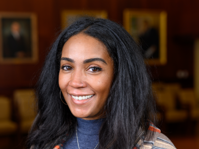 Headshot of Laneshia Conner, PhD, a Black woman in her 30s with long straight hair; she's smiling at the camera wearing salmon/blue/gray striped sweater and a blue turtle neck. In the blurred background is a maroon wall with large portraits.