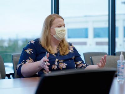 Alison Gibson, a white woman in her 30s, sits at a conference table with a wall of windows behind her. She has shoulder-length blonde hair, wearing a yellow procedure mask, and a navy blouse with big pink flowers. She's gesturing with her hands. 