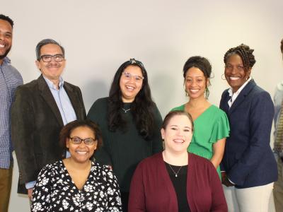 Photo of 8 adults of mixed races and skin tones. 6 adults are standing and two are sitting. They're in front of a white wall. Everyone is dressed in business casual clothes and smiling at the camera. 