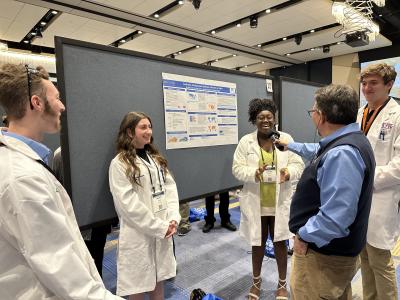 Four high school students--two white boys, one white girl, and one Black girl--stand in white lab coats in front of a research poster pinned to a board. The Black female student is speaking into a microphone held out by a middle-aged white male reporter, whose back is to the viewer. 