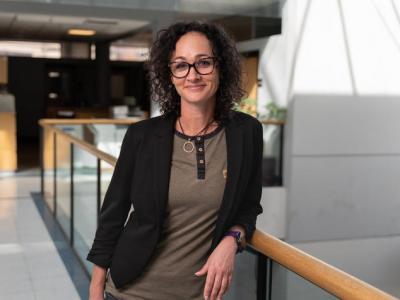 Photo of Sally Ellingson, PhD. She's a white woman in her 30s with very curly dark brown hair down to her shoulders and dark-rimmed glasses. She's wearing a dark brown blazer and a light brown shirt with dark trim. She's standing in a brightly lit building and leaning against a wooden guard rail. She's smiling at the camera. 
