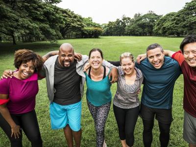 Group of six adults of various ages stand in a park-like setting with their arms around each others' shoulders. They're smiling at the camera and wearing workout clothes. 