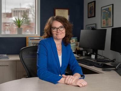 Portrait of Erin Haynes sitting at her desk in her office. She's wearing a royal blue blazer with a white shirt beneath and her hands are folded together on the table. She has reddish-brown wavy hair just past her shoulders and she's wearing glasses. Behind her is a dark blue wall with a window, and to the right  are her computer monitors and framed pictures on a white wall. 