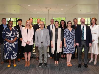 A diverse group of 16 faculty members stand together. They are all dressed professionally and behind them is a wall of windows. 