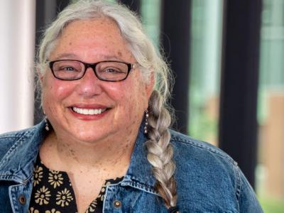 Linda Dwoskin is a white woman with long gray hair in a braid over her shoulder. She's smiling at the camera, wearing black-rimmed glasses, a black blouse with with flowers, and a denim jacket. A wall of windows is behind her. 