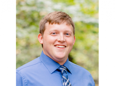 Headshot of Matthew Coleman, a young white man with short, light brown hair. He's outdoors, with trees blurred in the background, and he's wearing a light blue collared shirt with a blue and gray striped tie. 