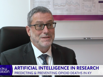 Screenshot of interview with Jeff Talbert, PhD. He's a white, middle-aged man sitting in an office chair wearing a gray suit, white shirt, and dark tie with black-rimmed rectangular glasses and short gray hair. There's a white board behind him and the bottom of a research poster is visible. At the bottom of the screen is a text banner that says "Kentucky Edition. Artificial Intelligence in Research: Predicting & Preventing Opioid Deaths in KY"