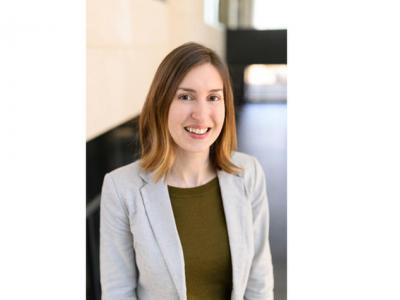 Photo of Laurie McLouth, a young white woman with straight, shoulder-length light brown hair. She's wearing a light gray blazer with an army green shirt, and smiling at the camera. Behind her is a beige wall and a window. 