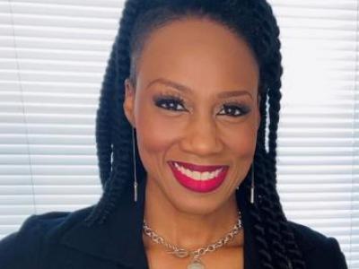 Headshot of Ketrell McWhorter, a young Black woman with shoulder-length twists, half pulled up into a high bun. She's smiling at the camera, wearing red lipstick, a black suit jacket, and there's a window with blinds behind her. 