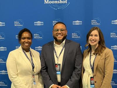 Three adults stand in front of a royal blue backdrop that says "Biden Cancer Moonshot" at the top. On the left is a Black woman wearing a cream colored blazer and dark trousers; in the center is a Black man with glasses, gray blazer, cream shirt, and plaid trousers; and on the left is a white woman in a tan blazer, cream blouse, and dark trousers. They're each smiling and wearing an event lanyard. 