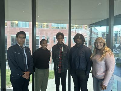 Five undergradute students stand side by side in front of a floor-to-ceiling window. 