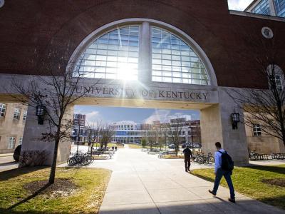 A brick and glass pedestrian bridge with a large half-moon window, under which are the words University of Kentucky. Light is shining through the window, creating a small sun burst. Two individuals are walking under the pedestrian bridge. 