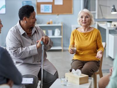 Two older women, one Black and one white, sit beside one another in a discussion circle in an office. The Black woman has short cropped hair and is holding her cane. The white woman has chin-length white hair and is holding a glass of water. We see the shoulders and hands of other members of the group discussion who are out of frame. 