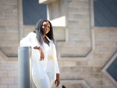 A young Black woman dressed in a white suit with gold belt stands and leans her right elbow on a gray bollard. She has chest-length black hair and is smiling at the camera. In the background is a very large stone sculpture of the UK inisignia. 