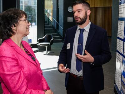 A young white man in a dark blue suit and navy tie presents a research poster to NIH director Monica Bertagnolli, a middle-aged white woman wearing a pink suit. 