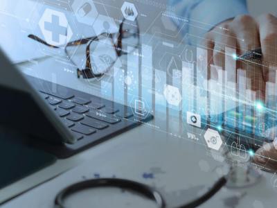 Stock image showing hands working at a laptop, and a stethoscope is on the desk. The image is overlaid with digital imagery of bar graphs and iconography. 