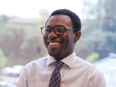 A young Black man is giving a big smile and looking slightly off camera. He has short hair, black rimmed glasses, and is wearing a cream colored collared shirt with a burgundy tie. He's standing in front of a window, with foliage visible behind him. 