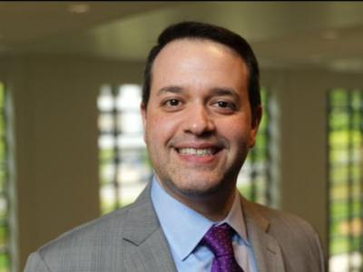 Headshot of Bill Stoops, a middle-aged white man whit short dark hair. He's smiling at the camera, wearing a light gray suit, light blue shirt, and dark purple tie. There's a wall of tall windows behind him. 