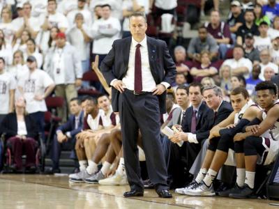 Photo of Billy Kennedy on the sideline of a basketball game. He's wearing a dark suit, white shirt, and maroon tie. His hands are at his hips, and basketball players are seated behind him. 