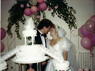 Wedding photo of a young white couple. They're standing behind a muiti-tiered wedding cake, with their foreheads touching. An arch of greenery and pink balloons is above them. 
