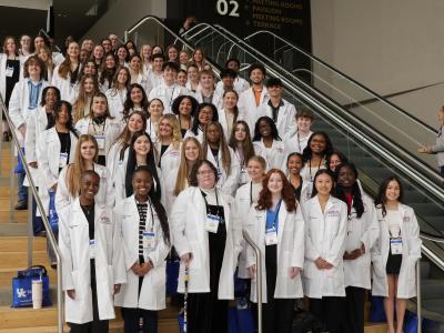 66 high school students in white lab coats stand on a large staircase of a conference center. 