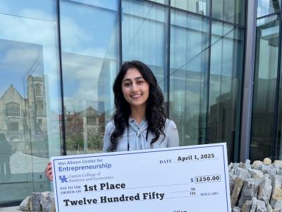 A young woman with long, wavy, dark hair stands holding a giant ceremonial check that says "First Place: $1250". She's standing outside on a paved walkway with a glass building behind her. 