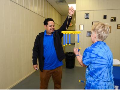 A young male community health worker holds up a coordination game for an older community member. The health worker is a young Hispanic man with dark hair, wearing a blue polo, black jacket and brown trousers. He's smiling as he dangles the game in front of a an older white woman wearing a blue tie dyed t-shirt. 