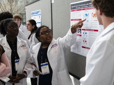 High school students in white lab coats gather around a research poster; one of them is pointing at the poster and speaking. 