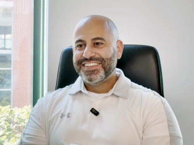 A smiling man sitting in a office chair; he's wearing a white polo shirt and has a bald head and light beard. Behind him is a white wall and a window. 