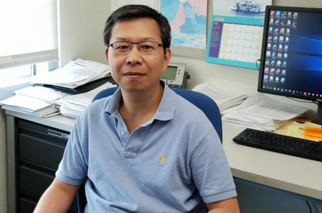 Dr. Dr. Qing-Bai She sits facing the camera with his back to his desk and computer screen. He is wearing a pale blue polo shirt and glasses. 