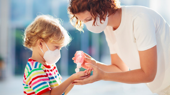 A parent in a white t-shirt squeezes hand sanitizer into the hands of a blond child wearing a rainbow striped shirt. Both people are wearing masks.