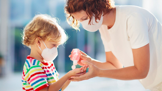 A parent in a white t-shirt squeezes hand sanitizer into the hands of a blond child wearing a rainbow striped shirt. Both people are wearing masks. 